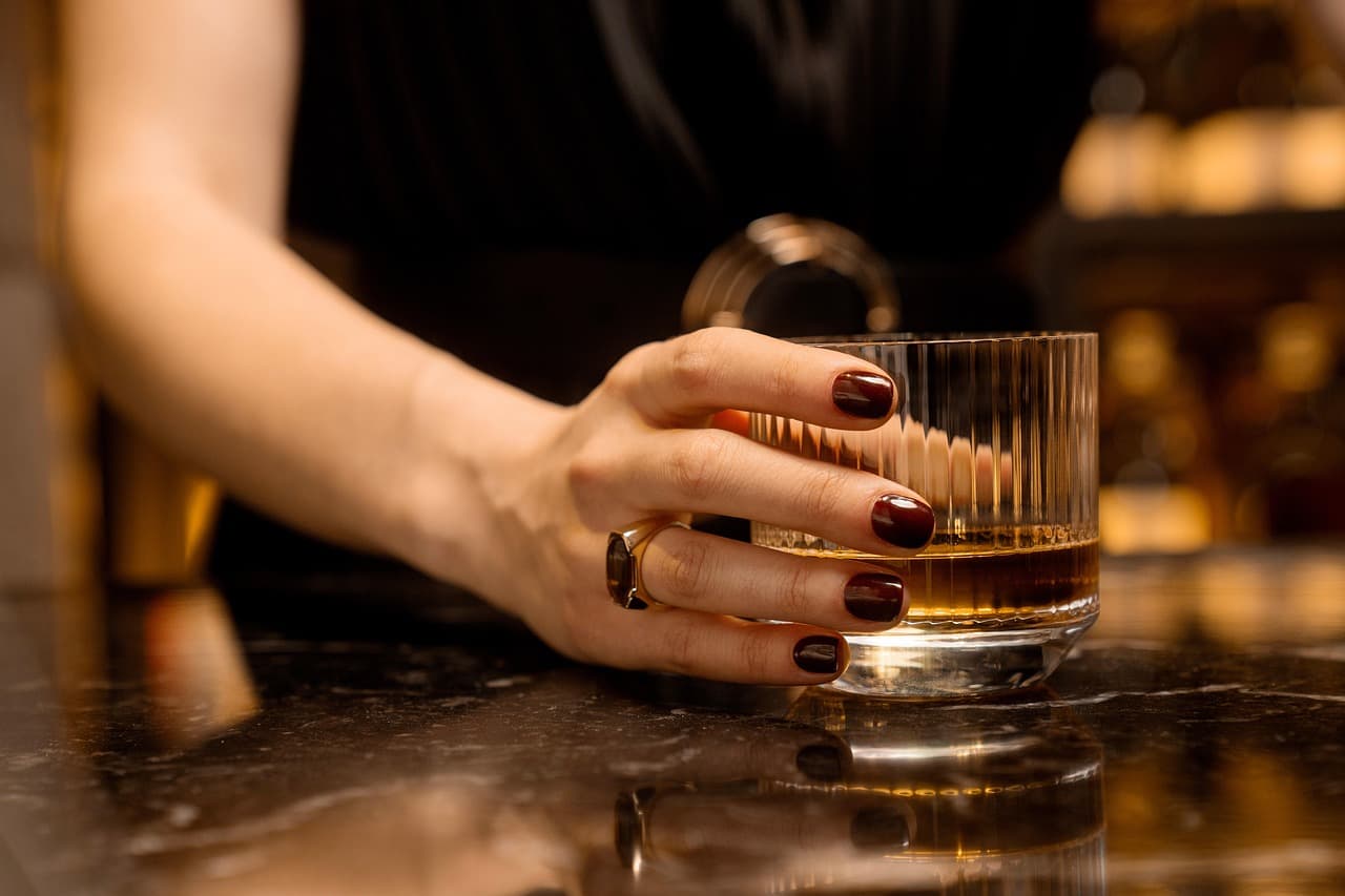 A hand with a deep burgundy manicure and a gold ring resting on a glass
